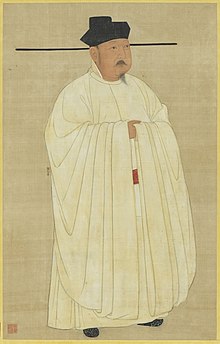 A short man in heavy white robes, wearing a black hat with long horizontal protrusions coming from the bottom of the hat. The man has a small, pointed beard and a small mustache.