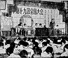 Group of Zengakuren leaders sitting on a stage, with a Zengakuren leader clad in black making a speech in front of a crowd of students.