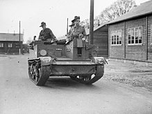 Black and white photo of three men wearing military uniforms standing in a tracked military vehicle which is on the road in a camp