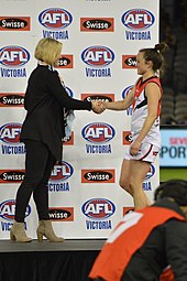 Pearce shaking hands with a woman onstage as she is presented with a medallion