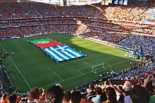 View from the stand of the pitch and opposite stands, with large flags covering a large part of the playing surface, held aloft by a number of flag-bearers