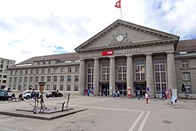 Farbfotografie von einem dreigeschossigen Gebäude mit langen Rundbogenfenstern im Säuleneingang. Im Giebelfeld ist die Uhr von zwei Figuren umrandet. Auf dem Dach weht die Schweizer Flagge.
