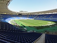 Blick von den leeren Zuschauerrängen einer schattigen Kurve im Olympiastadion Rom aufs Fußballfeld bei blauem Himmel