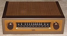 A small rectangular brown box with a metallic coppery top on a beige carpet. On its front are two dials and a tuner with a range between 88 and 108