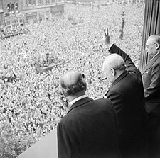 A balding man with a cigar in his mouth waves from a balcony to large crowds below him that fill the square.