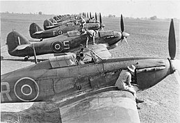 Side view of eleven World War II fighter planes parked in a very large field. Soldiers in khaki uniform, wearing shorts, climb atop or sit inside all of them making repairs.