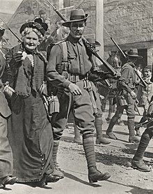 Troops marching through a city street lined with a crowd of people. In the foreground is a soldier and beside him is an older women.
