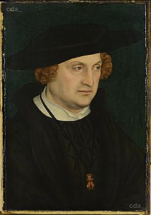 Middle-aged man in black, with a large black hat and light brown hair.