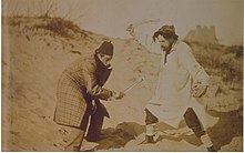 Sepia-Fotografie von zwei kostümierten Männern in einer Dünenlandschaft, die miteinander zu fechten scheinen. Der linke trägt einen karierten Mantel und einen Fez, der rechte einen hellen Kittel und eine Mütze. In den Händen halten sie Knochen und einen Totenschädel.