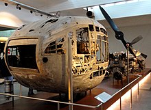 A dilapidated egg-shaped streamlined nacelle on display in a museum. The pointed end is towards the camera and has a large adjustable rectangular vent.