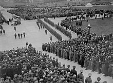 Black and white photo: Crowds of people and a ranked military guard gather to watch four men in ceremonial uniform approach a set of stone steps.