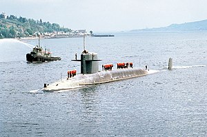 Angled view of gray submarine in river steaming towards camera. Personnel wearing high-visibility apparel are standing atop the submarine, while a tugboat spraying water is visible at the submarine's starboard side.