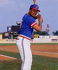 A man wearing a blue baseball jersey with red and white trim, a blue cap with a white "N" on the center, and white pants stands on a baseball field posed with bat, ready to swing.