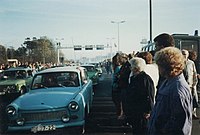 East German Trabant cars driving between dense crowds of people. Metal gantries over the road and a border watchtower are visible in the background.