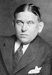 A black and white photographic portrait of critic H. L. Mencken. His hair is parted in the middle, and he appears to be leaning on his left arm. He is wearing a dark tie and a dark suit with peak lapels. A white handkerchief is visible in his suit pocket.