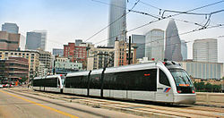 Two-car train running southbound towards Fannin South station on the METRORail Red Line. The skyscrapers of downtown Houston can be seen in the background, and overhead wires are visible to provide power to the train.