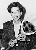 A woman looking toward the camera with a tennis racket held diagonally in front of her.