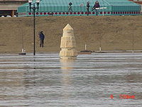 Water from a river completely surrounds a stone obelisk. In the background are several people with news cameras. A building with a green roof is seen behind them. At the bottom right of the image is the camera's time: "6 7:59 am".