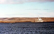 A warship at anchor. In the background is a flat, treeless landscape.
