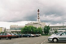 Frontale Farbfotografie eines Industriegebäudes mit drei dicken Rohren, die in der Höhe verbunden sind. Links steht auf dem Dach „Valgykla“. Im Vordergrund stehen auf einem Parkplatz Autos.