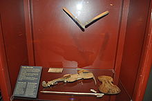 Photograph of a display case with drumsticks suspended in the air as though poised over a drum along with the top and bottom covers of a violin with a violin bow