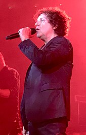 A man with a dark curly hair wearing a dark jacket, singing into a microphone