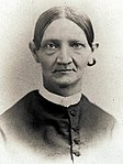 old sepia photograph of Lucy Smith Millikin, taken between 1850 and 1900. The older woman has tightly pulled hair, prominent ears, and slight shoulders. She is dressed in a dark dress with a white collar.