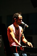 A man facing right is performing a keyboard with a microphone in front of him