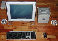 The G4 Cube sits besides a large, flatscreen monitor in matching grey. Flanking the monitor and Cube are round speakers clad in translucent plastic, and a keyboard and mouse.