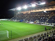 An evening game at a football stadium. The main stand on the right is filled with spectators and the empty pitch is being prepared for playing on the left