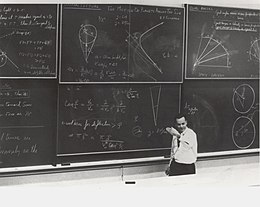 Feynman standing before a large blackboard with chalk writing all over it