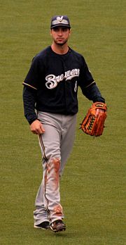 A man wearing a navy blue jersey with "Brewers" on the front in white, gray pants, navy blue cap with a white "M", and outfielder's glove on his left hand walking on a grassy field