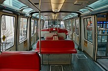 The interior of a monorail train with a slightly curved ceiling, large windows and doors on both sides, and red seating in the middle that face towards the camera and the sides
