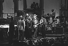The Band standing onstage, encircling Dylan around a microphone