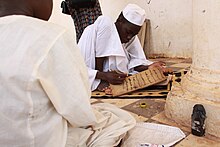 A black man in a cream coloured robe watches a boy in a white robe and white cap write in Arabic on a flat piece of wood. They are both sitting on a tiled floor near a white column.