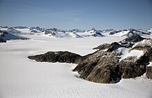 A distance shot near Juneau, Alaska. The area is covered in untouched white snow with mountain peaks in the distance. To the right of the image, nearer the photographer, is an uncovered outcrop of rocks.