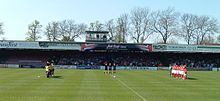 One of the stands of the Bootham Crescent association football ground, with supporters sitting down and players standing on a grass field below