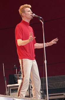 A middle-aged white man with orange hair, a red shirt and striped pants, singing in front of a microphone on stage