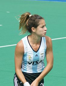 Giselle Kañevsky trägt einen schwarzen Rock und das blau-weiß-gestreifte argentinische Nationaltrikot. Die Haltung der beiden Hände deutet darauf hin, dass sie ihren Schläger führt, dieser ist aber nicht im Bild.