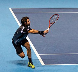 A man wearing a black shirt, black shorts, and neon yellow shoes staggers to his right to return a tennis ball as he holds his red tennis racket in front of him