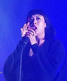 Alisa Xayalith wearing dark clothes, singing into a microphone onstage