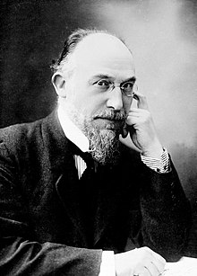Elderly white man, balding, with neat moustache and beard, in formal daywear with wing collar and dark necktie. He is wearing pince-nez glasses