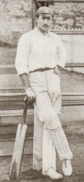 A posed black and white photograph of a cricketer on the edge of a cricket field. He is leaning against the post of a scoreboard with his legs crossed and bat leant in one hand.