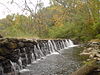 Several streams of water fall over a dam made of large rocks under autumn leaves