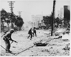 U.S. Marines engaged in urban warfare during the battle for Seoul in late September 1950. The American soldiers are carrying M1 Garand semi-automatic rifles and Browning Automatics. On the street are Korean civilians who died in the battle. In the distance are M4 Sherman tanks.