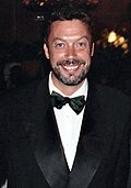 A bearded man, wearing a white shirt, black bowtie, and black jacket, smiles at the camera.