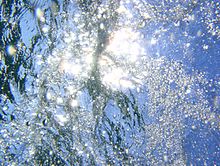 Unterwasser-Fotografie einer Wasseroberfläche aus etwa 20 cm Tiefe, in Richtung einer Lichtquelle. Die Wasseroberfläche ist gewellt, die Wellen brechen die Lichtstrahlen, sodass nicht erkennbar ist, woher das Licht genau kommt. Luftblasen steigen zur Oberfläche hin auf.