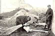 Two men look at a propeller and gear assembly lying on the ground. One is stooping over it, inspecting it closely; the other leans with his back against a whitewashed wall on the right. Buildings are visible in the background.