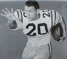 Black and white posed shot of Cannon from the thighs up, in the stiff-arm position, cradling a football, wearing pads and a jersey number 20, helmetless