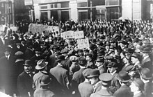 Black and white photograph of a large crowd of people, a few holding signs above the crowd, displaying IWW acronyms and slogans.